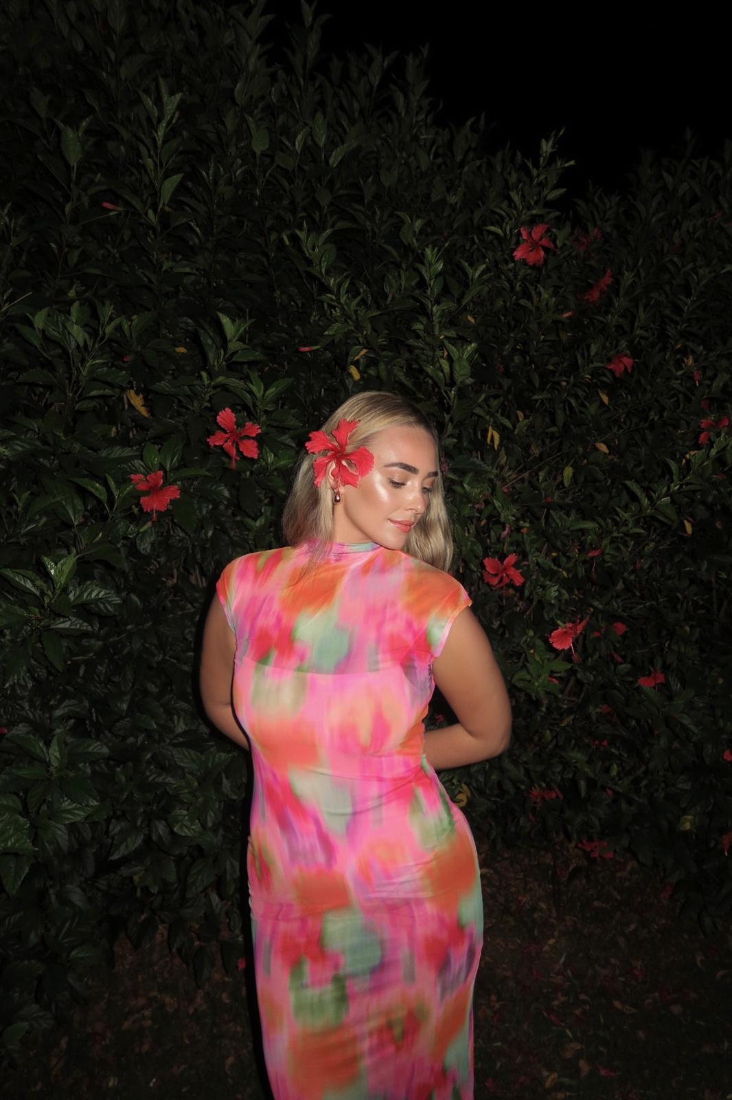 Bailey wearing a colorful floral dress with a hibiscus flower in her hair, standing against tropical greenery